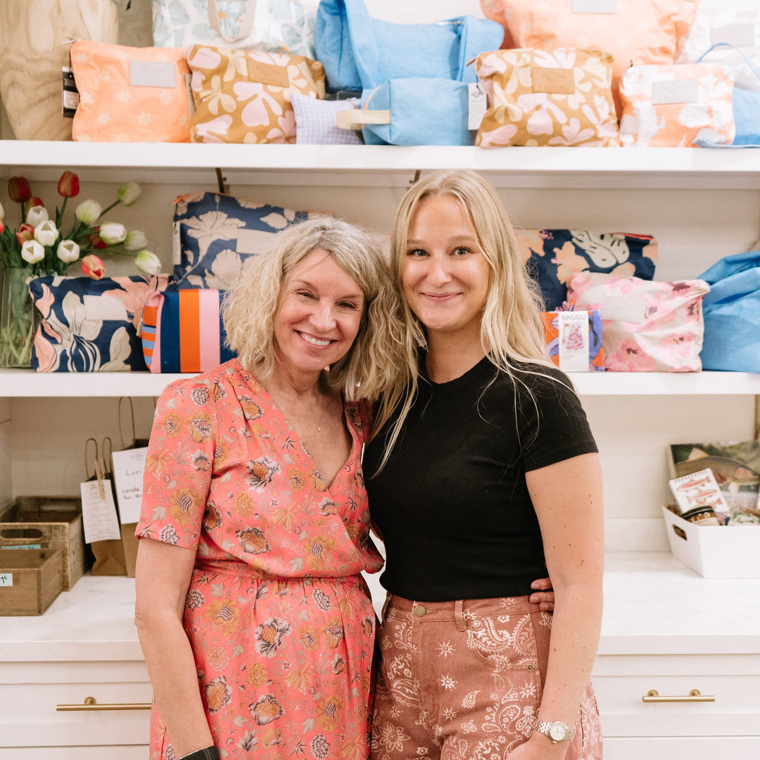 Two women standing together in a bright home or boutique setting, with shelves behind them filled with colorful patterned pillows and home decor. The woman on the left wears a coral floral dress, and the woman on the right wears a black top and rust-colored patterned pants.