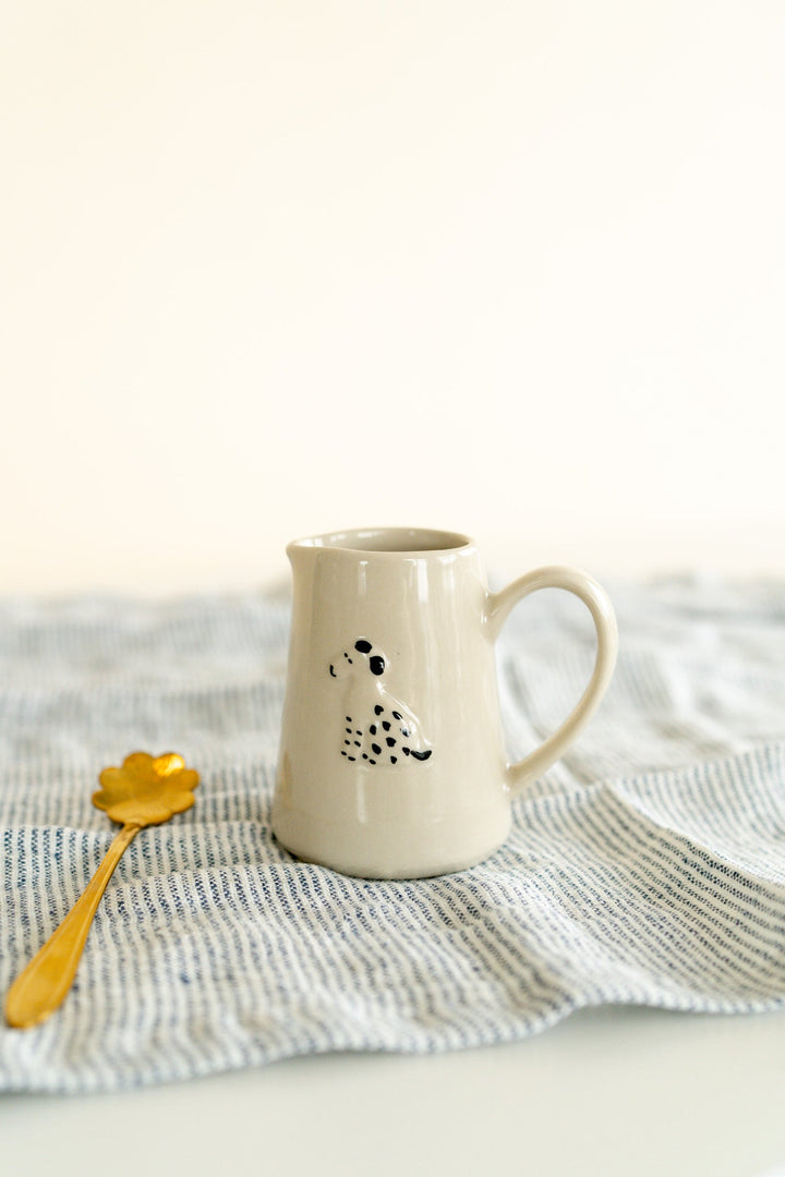 Dog Mini Creamer Pitcher - A petite pitcher with a painted black and white dog sits on a striped towel next to a gold spoon